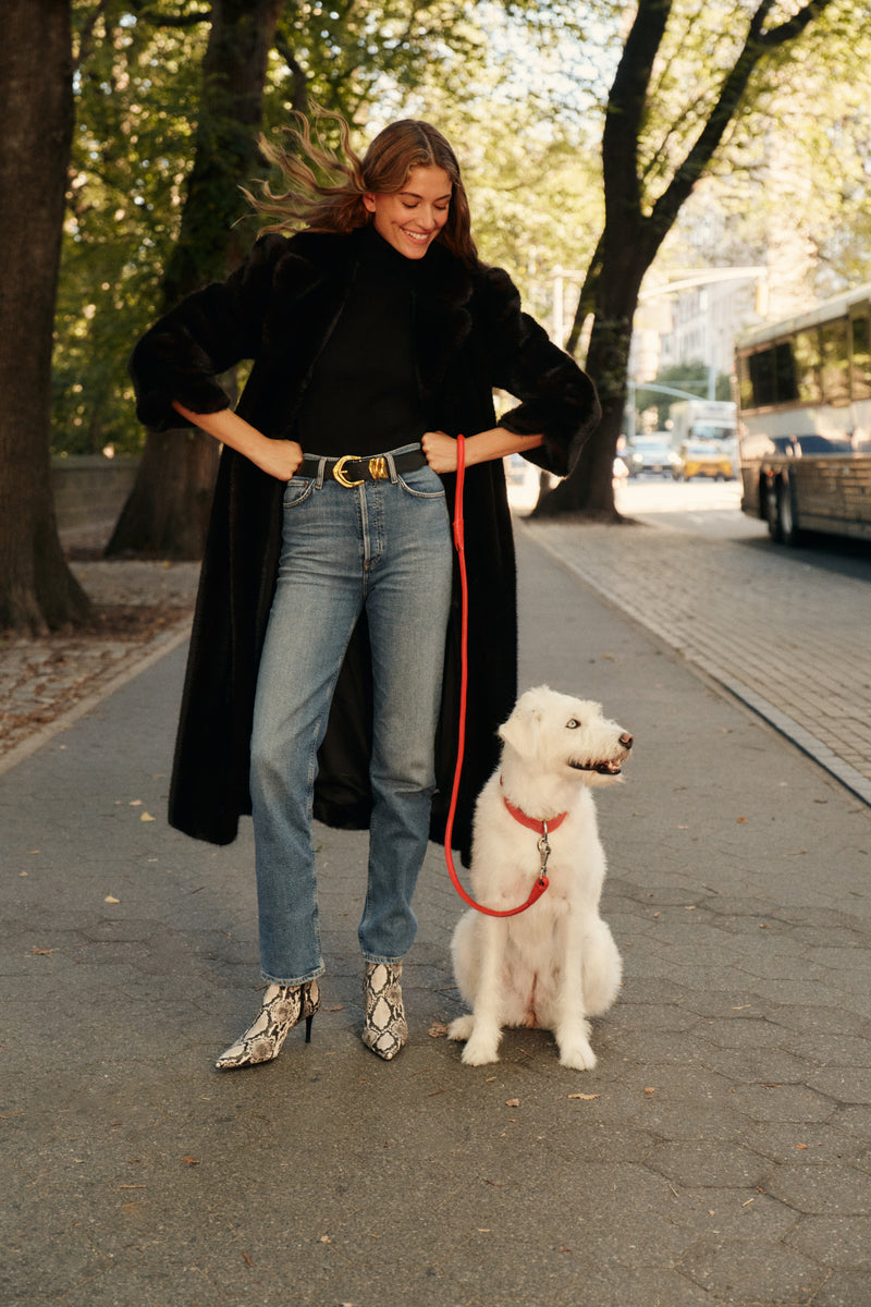 A woman walks a dog with a red leash while wearing a full-length fur coat, a black turtleneck, a pair of blue jeans, and a pair of snakeskin boots.