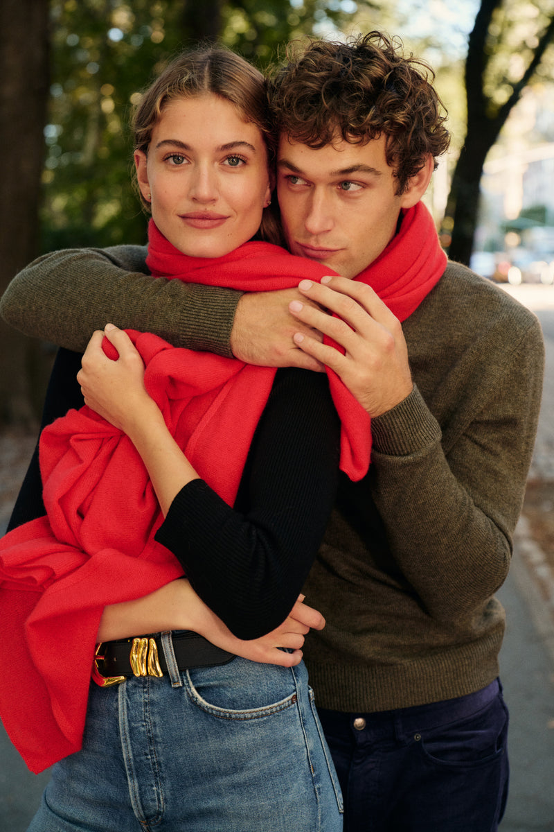 On the left, a woman stands wearing a red scarf, a black turtleneck, and blue jeans. On the right, a man stands with his arm around the woman while wearing an olive green sweater and dark blue jeans.