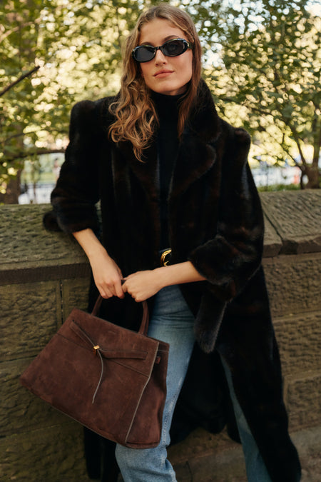 A woman leans on a city park bridge wearing a full-length fur coat, a black turtleneck, and a pair of blue jeans while carrying a brown suede tote bag and wearing sunglasses.