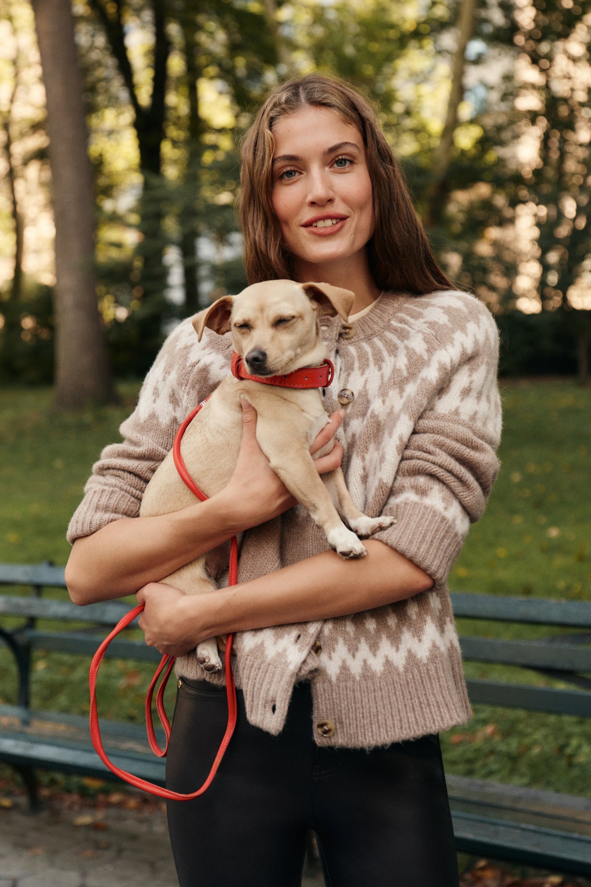 A woman holds a dog in a city park bench while wearing a fair isle patterned cardigan, black leather pants, and brown suede boots.
