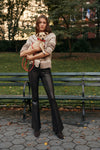 A woman holds a dog in a city park bench while wearing a fair isle patterned cardigan, black leather pants, and brown suede boots.