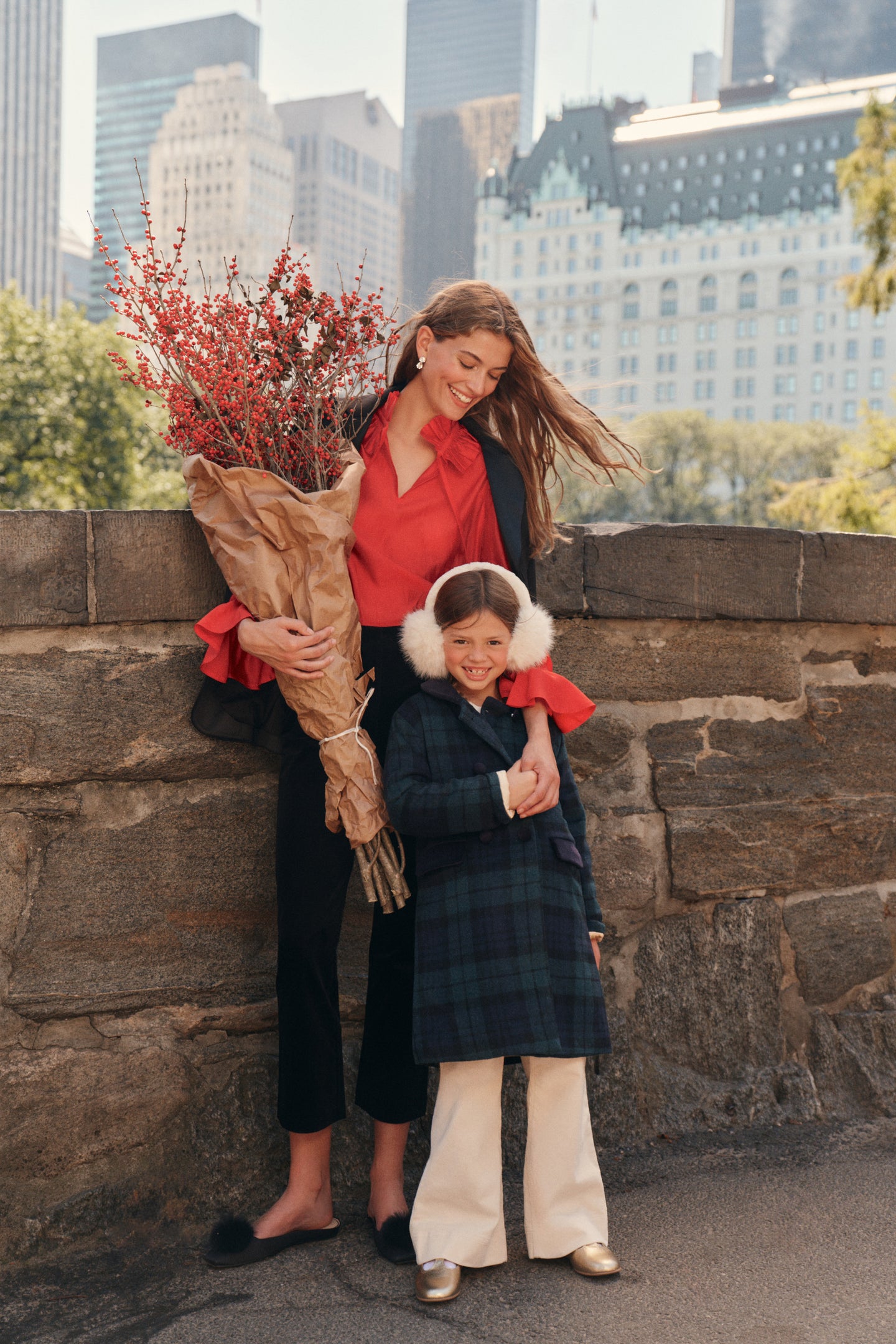 A woman stands on a city park bridge with a little girl. The woman wears a black blazer, a red blouse, black pants, and black flats while carrying a set of paper wrapped florals. The little girl wears a tartan patterned coat, cream pants, gold t-strap shoes, and ear muffs.