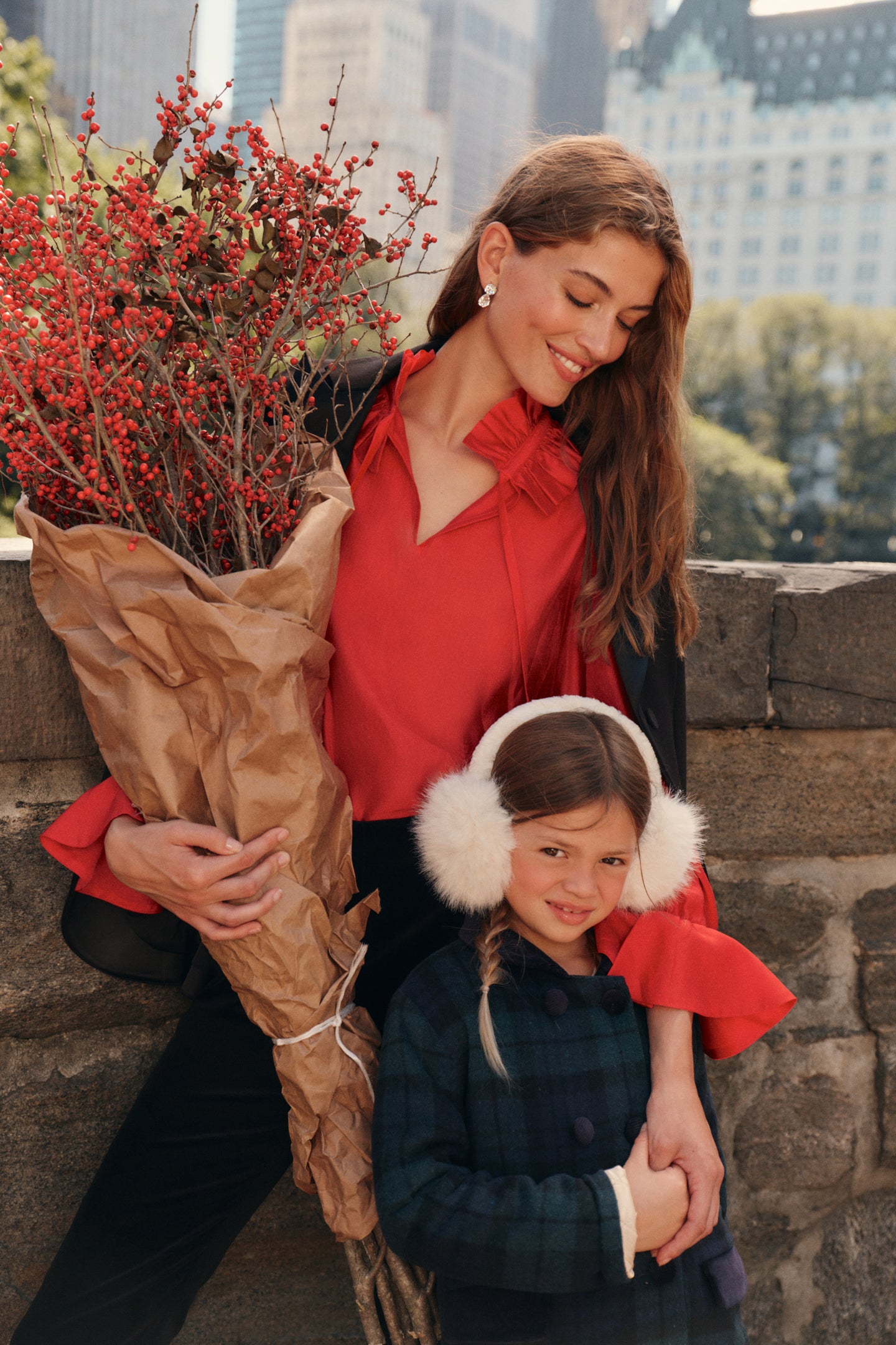 A woman stands on a city park bridge with a little girl. The woman wears a black blazer, a red blouse, and black pants while carrying a set of paper wrapped florals. The little girl wears a tartan patterned coat and ear muffs.