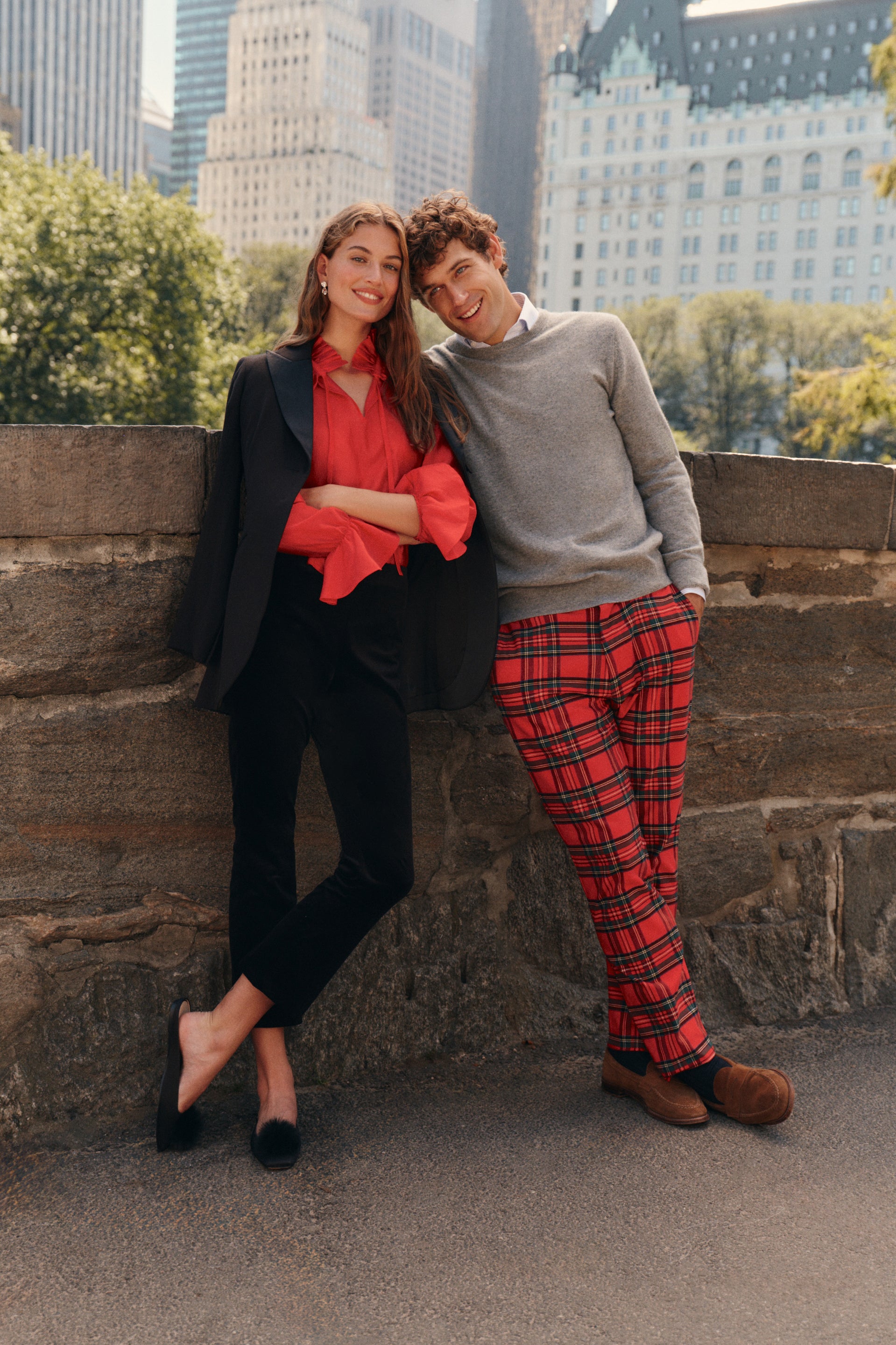 On the left, a woman stands wearing a red blouse, back pants, a black blazer, and black flats. On the right, a man wears a gray sweater over a white button down, red plaid trousers, and brown loafers.