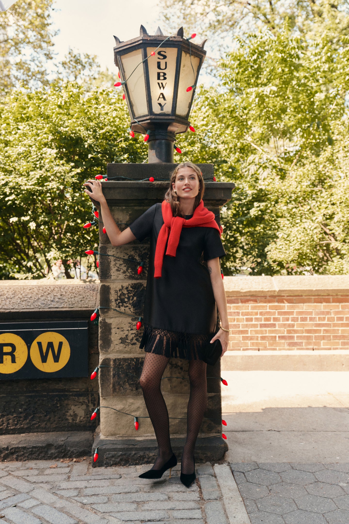 A woman wearing a black dupioni mini dress with tassels, a red sweater wrapped around her shoulders, polka dot tights, and black heels while carrying a black clutch in a city park.