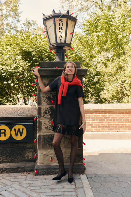 A woman wearing a black dupioni mini dress with tassels, a red sweater wrapped around her shoulders, polka dot tights, and black heels while carrying a black clutch in a city park.