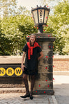 A woman wearing a black dupioni mini dress with tassels, a red sweater wrapped around her shoulders, polka dot tights, and black heels while carrying a black clutch in a city park.