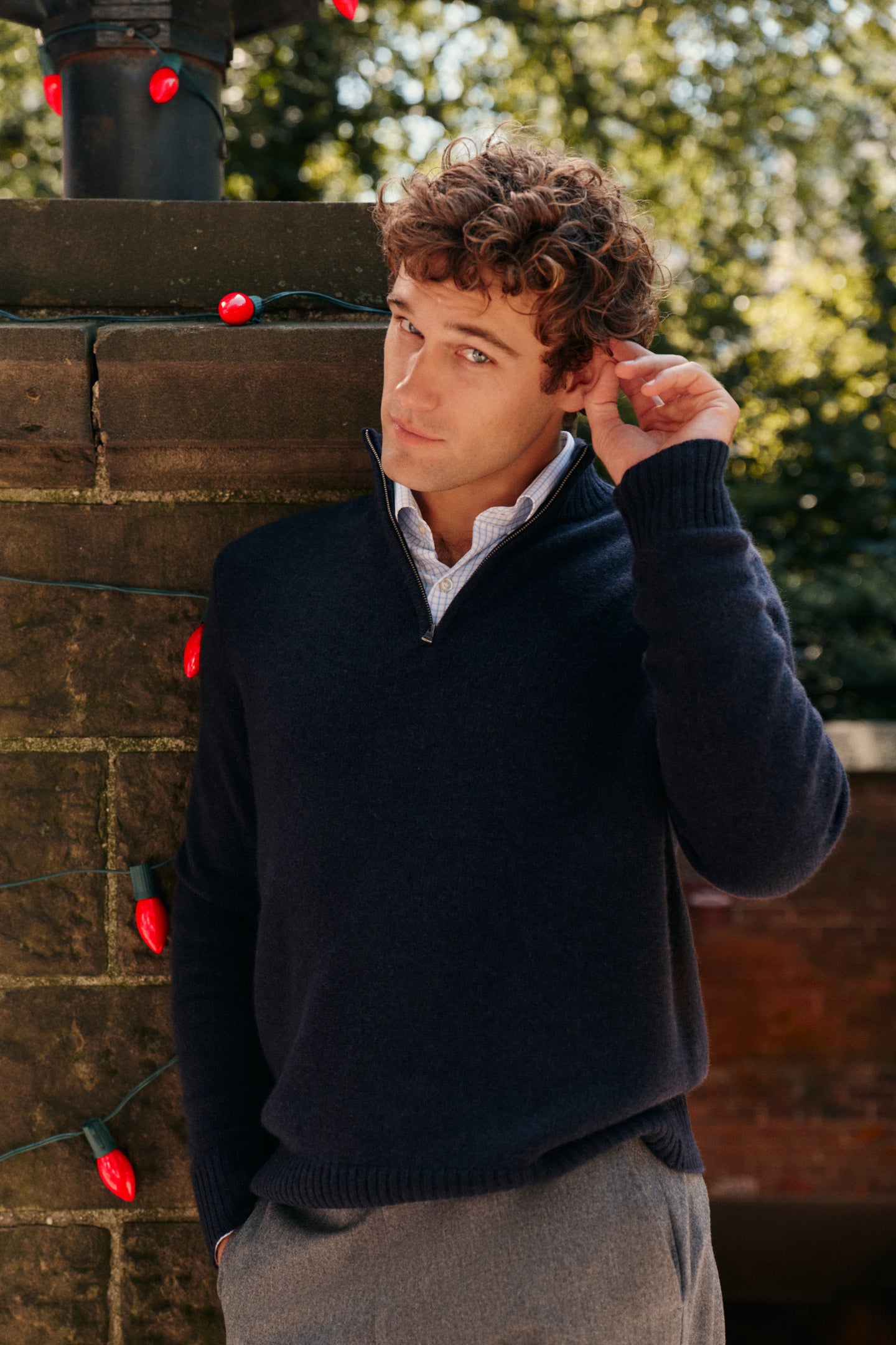 A man leans against a brick pillar, wearing a navy quarter zip and gray trousers.