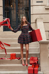 Young woman in a classic Baroque Tartan Bubble Liza Dress holds a red gift box, capturing an effortless, fun, high-end holiday vibe on brownstone steps.