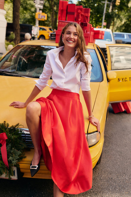 A woman leans against a yellow city cab wearing a white button down, a red dupioni midi skirt, and black heels.