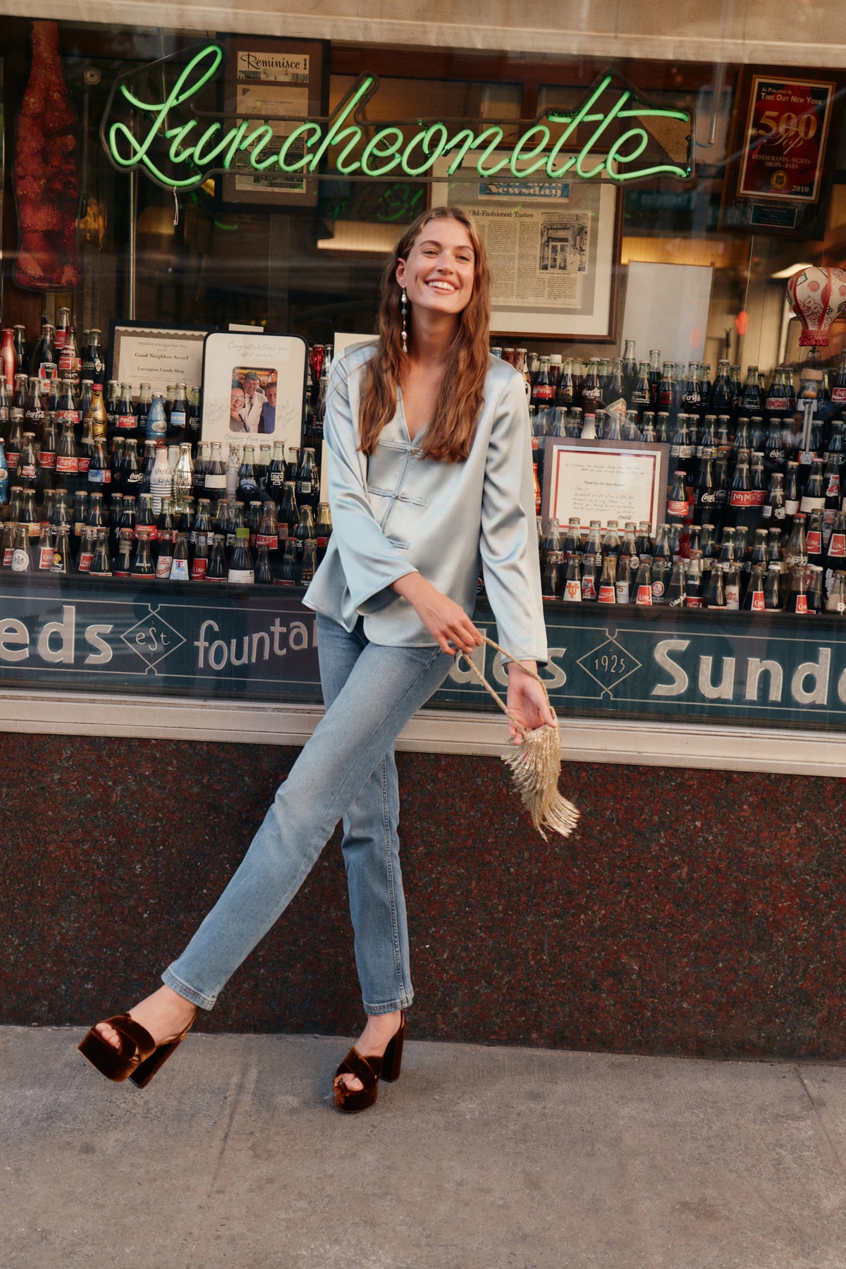 A woman wearing a blue satin top, blue jeans, and brown velvet heels while carrying a gold fringe purse.