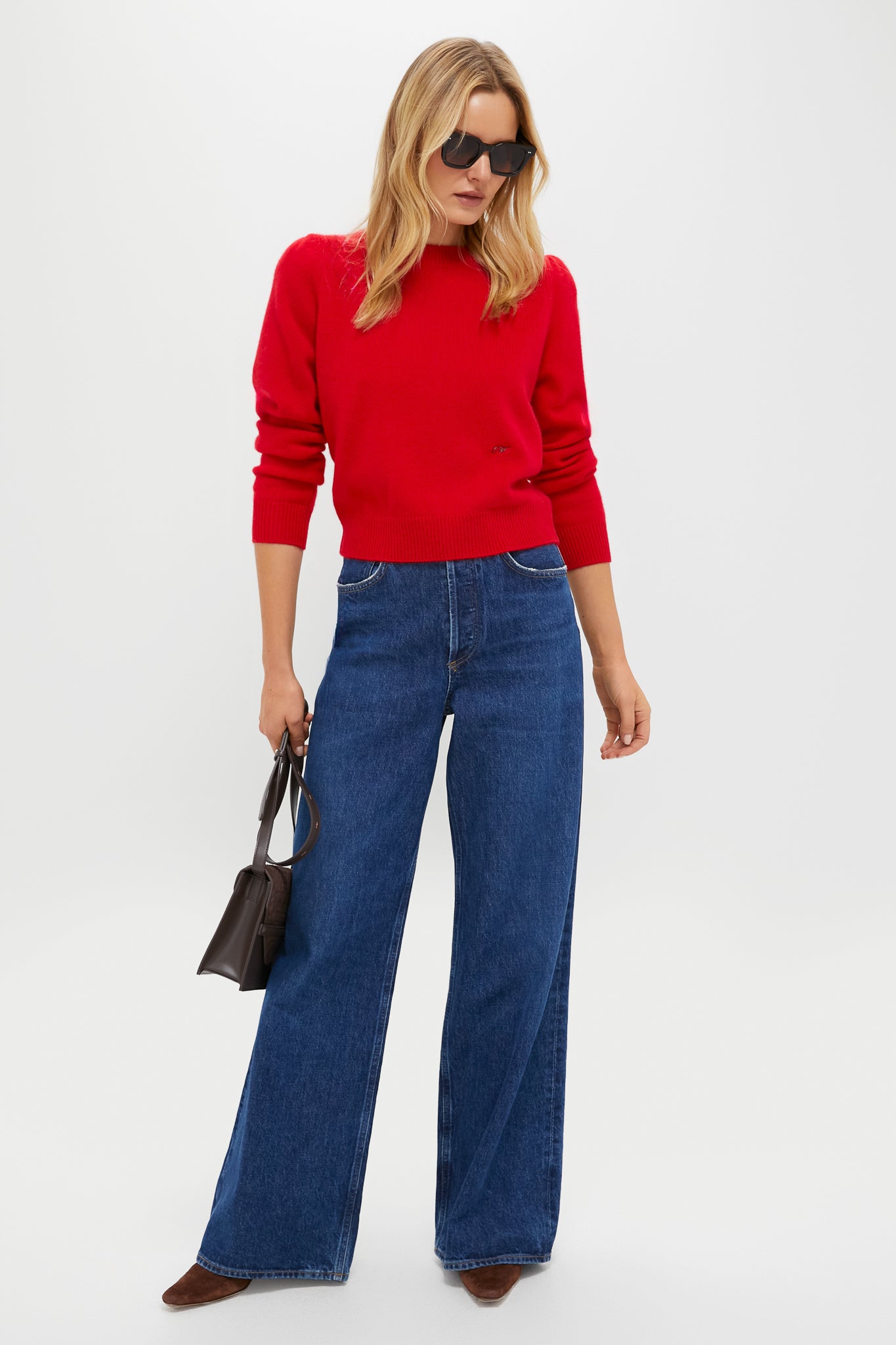 A woman in a Tuckernuck red cashmere crewneck sweater, wide-leg blue jeans, brown shoes, sunglasses, and carrying a small black handbag stands against a plain white background.