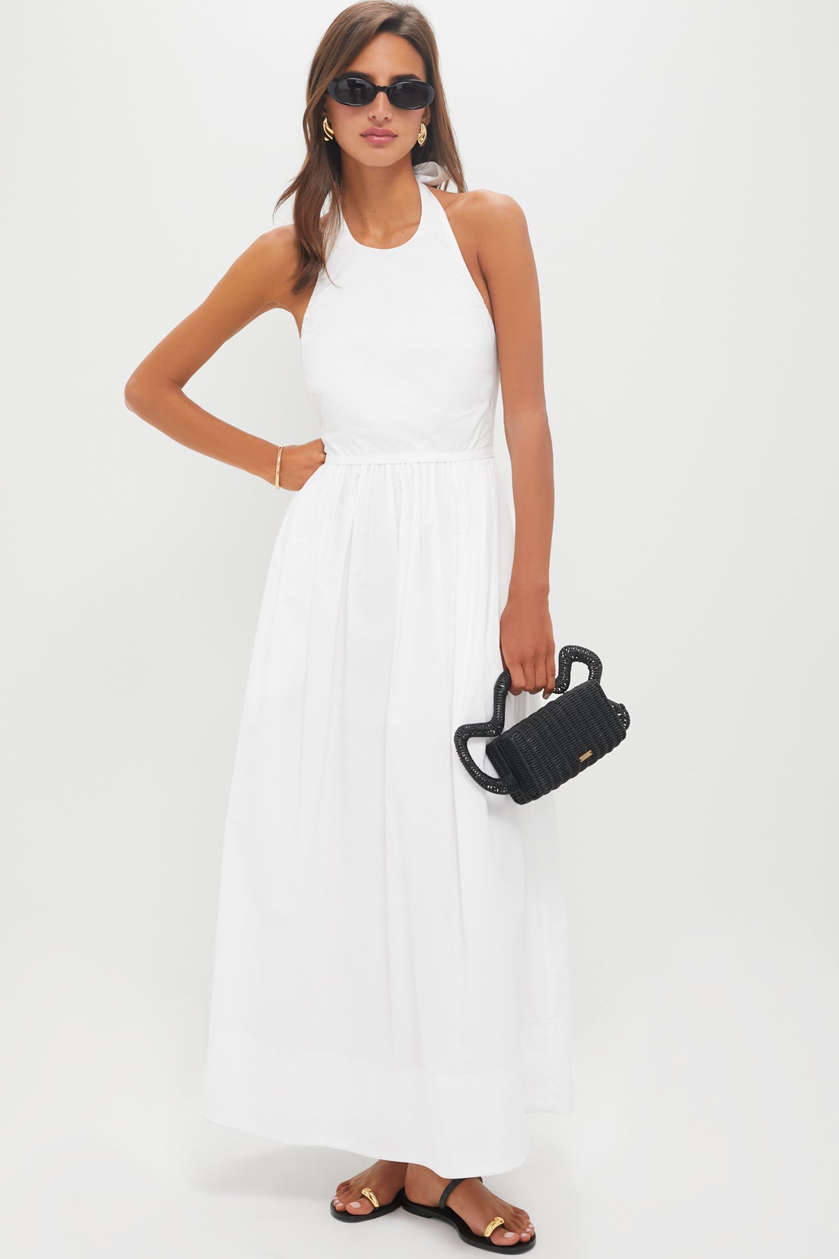 A woman in a sleeveless white 100% cotton Tuckernuck maxi summer dress, black sunglasses, black sandals, and holding a small black woven handbag stands against a plain white background.