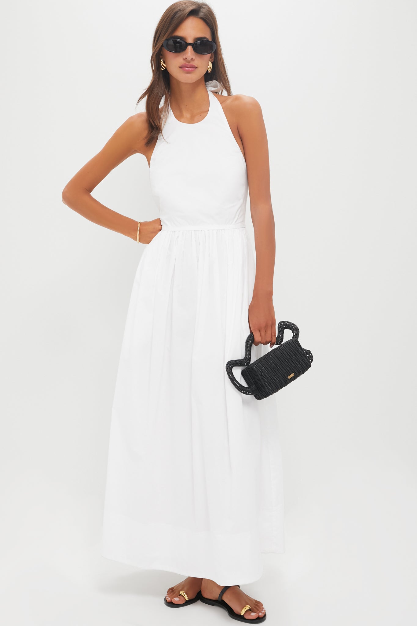 A woman in a sleeveless white 100% cotton Tuckernuck maxi summer dress, black sunglasses, black sandals, and holding a small black woven handbag stands against a plain white background.