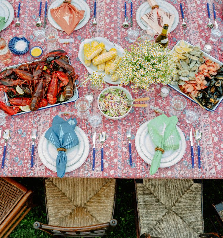 Overhead view of a summer table of corn, lobster, clams, and shrimp featuring a red tablecloth and colorful napkins. 