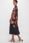 Woman modeling the Black Chenille Flowers Midi Dress with colorful floral print, scalloped trim, and blouson sleeves, styled with pink velvet mules for an effortless, high-end look.
