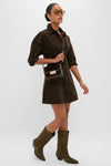 Woman wearing the Carob Luz Mini Dress, a classic corduroy shirt-dress with lantern sleeves, paired with tortoiseshell sunglasses, dark leather bag, and olive suede boots; effortlessly high-end.