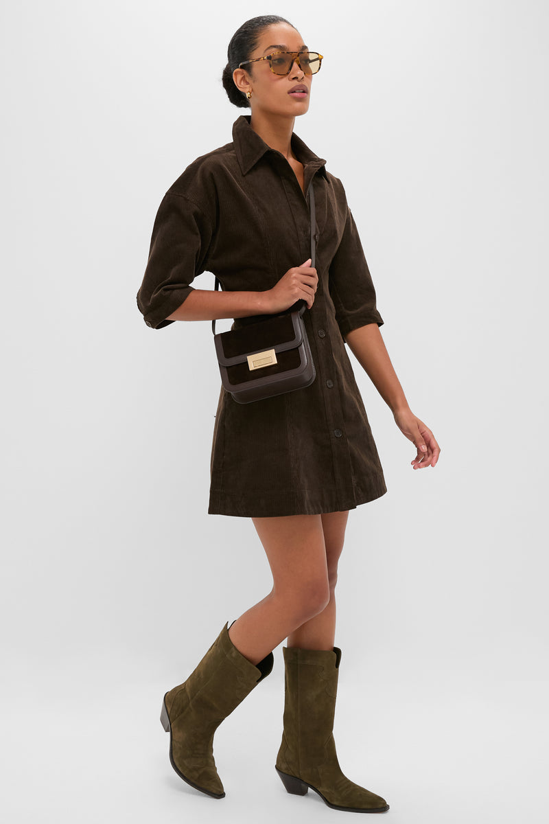 Woman wearing the Carob Luz Mini Dress, a classic corduroy shirt-dress with lantern sleeves, paired with tortoiseshell sunglasses, dark leather bag, and olive suede boots; effortlessly high-end.