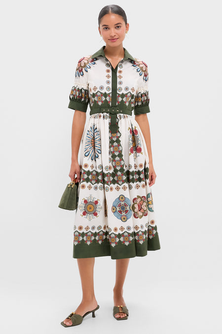 A woman wears the La DoubleJ Libertine Dress—white cotton poplin with green trim, floral and geometric prints—paired with a small green purse and green heeled sandals.