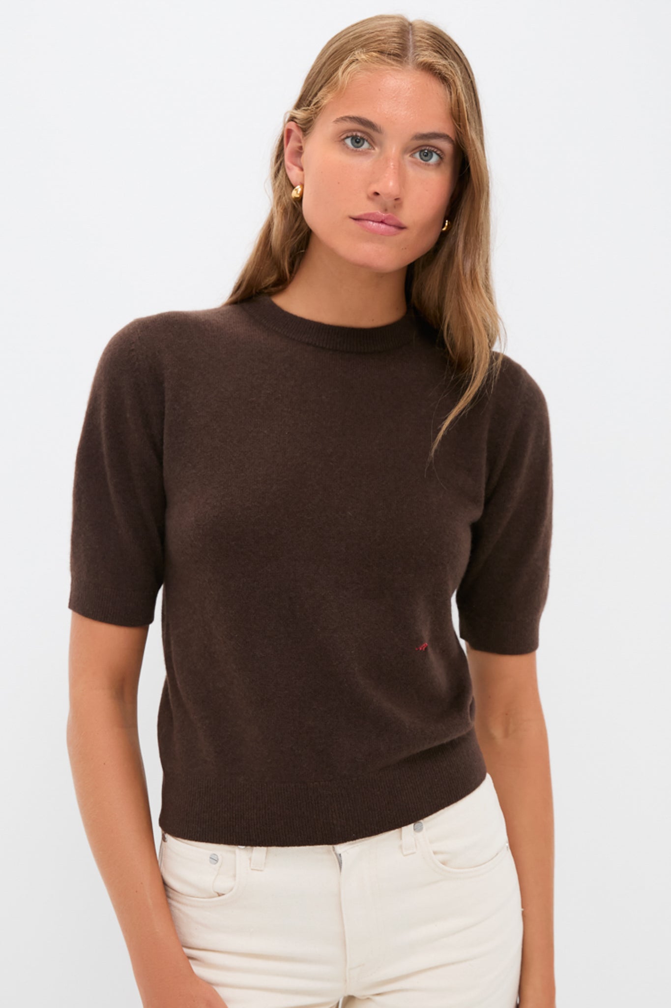 A woman wears a chocolate brown, short-sleeved Tuckernuck cashmere crewneck sweater with light-colored pants, standing against a plain white background.