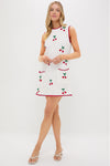 A woman models a Saylor sleeveless summer mini dress with red trim and cherry patterns, paired with white heeled sandals, against a plain white background.