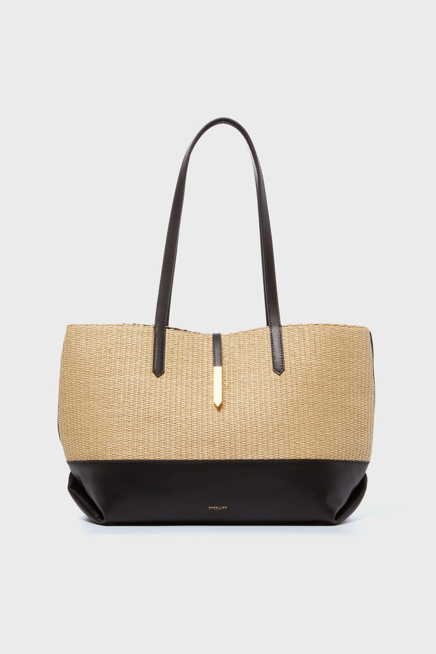 The Demellier woven raffia tote features a beige and black design, two dark handles, and a gold accent, highlighting the brand’s elegant handcrafted style.