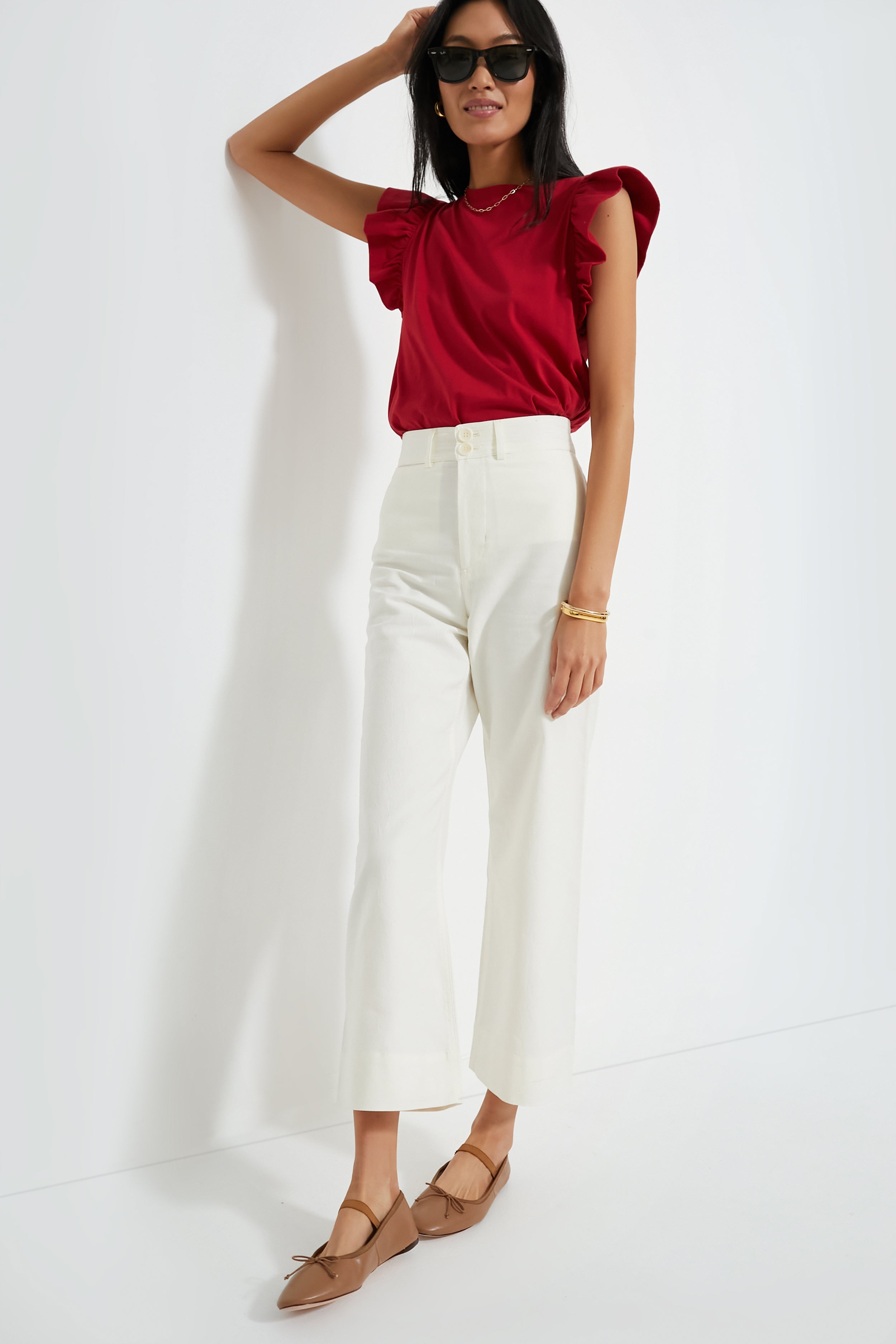 A woman in a Tuckernuck Red Kaia Flutter Sleeve Tee, white high-waisted pants, tan flats, and sunglasses stands confidently.