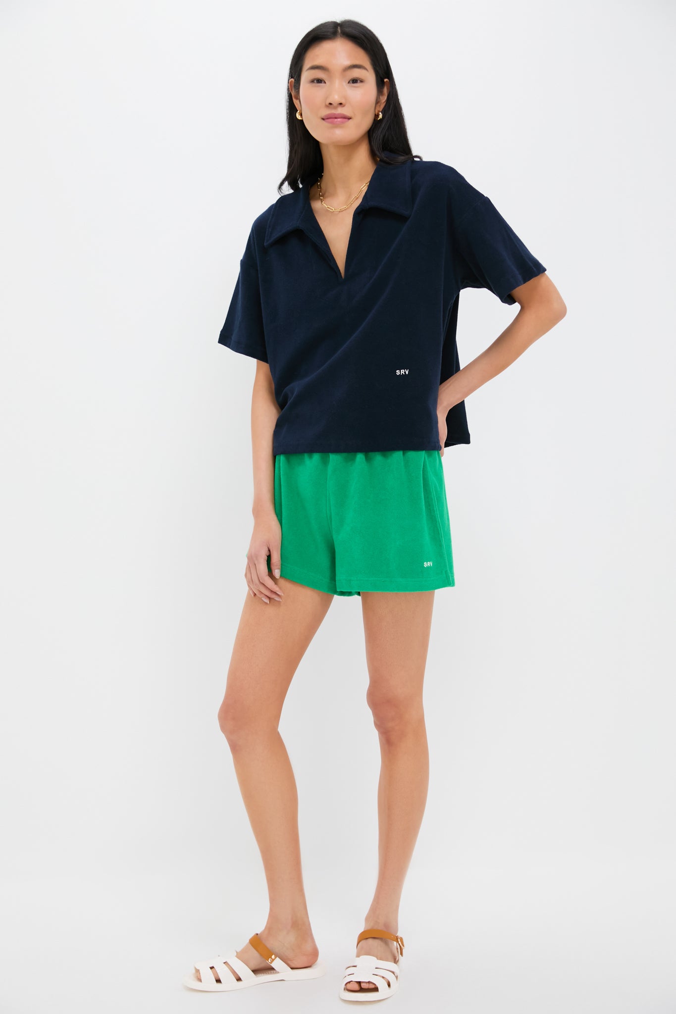 A woman models Tnuck Beach: a navy short-sleeve collared shirt, terrycloth shorts with an elastic waistband, and white sandals with tan accents—an effortless summer look.