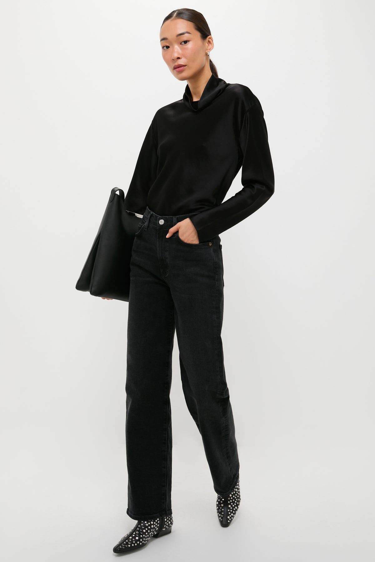 A woman in a LESET satin long sleeve top, black jeans, and studded boots stands with a large black bag against a white background.