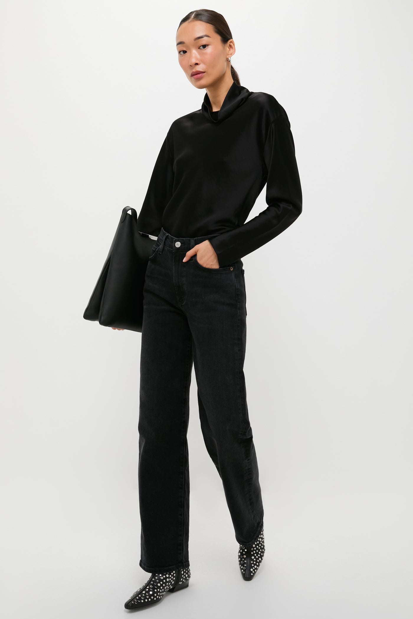 A woman in a LESET satin long sleeve top, black jeans, and studded boots stands with a large black bag against a white background.