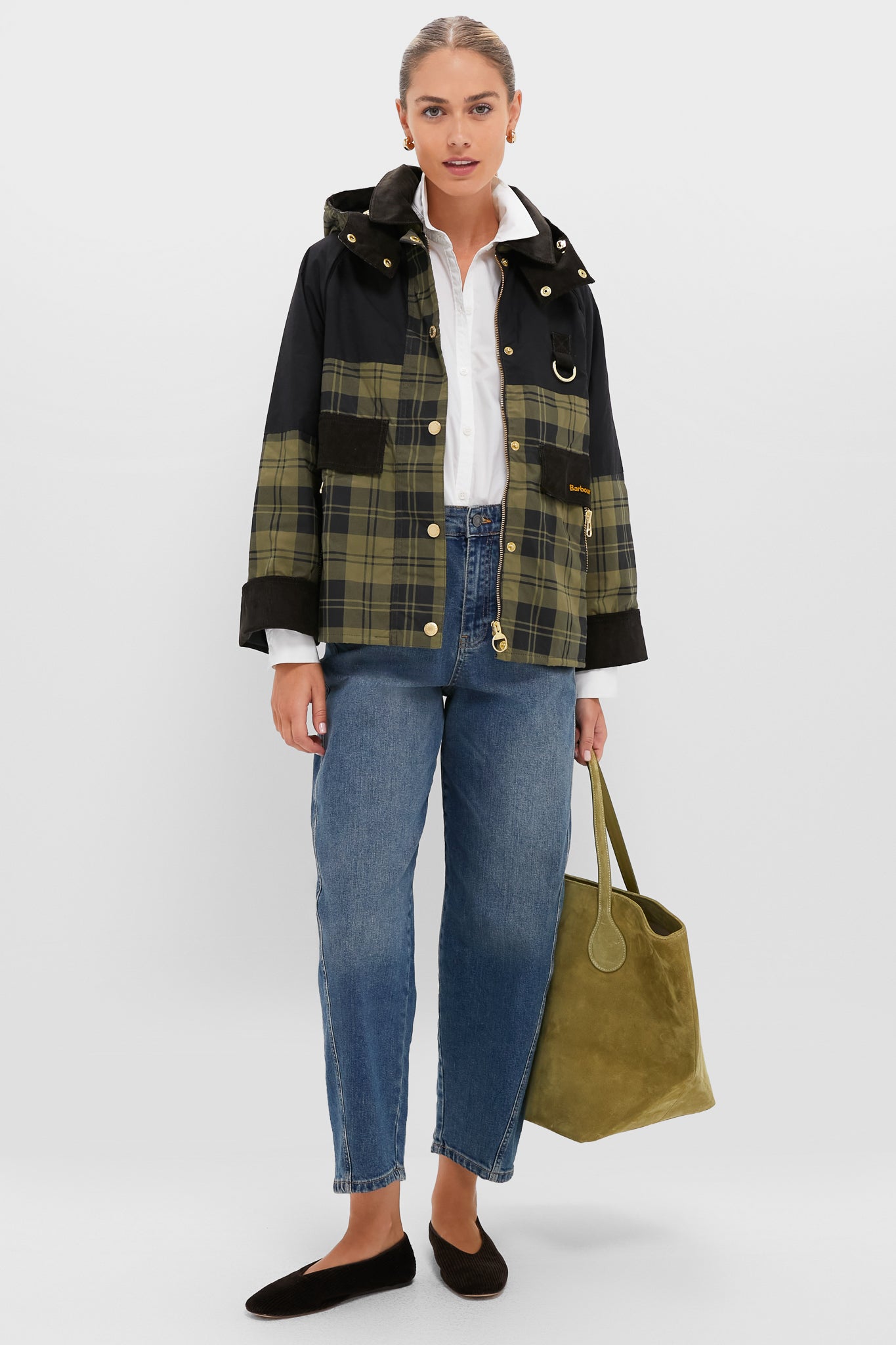 A woman stands wearing a Barbour black and green tartan jacket, white shirt, blue jeans, black flats, and holding a large olive green tote bag.