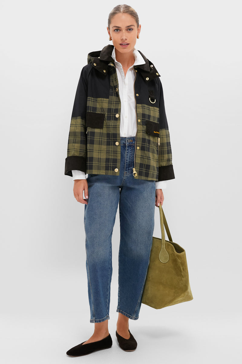 A woman stands wearing a Barbour black and green tartan jacket, white shirt, blue jeans, black flats, and holding a large olive green tote bag.