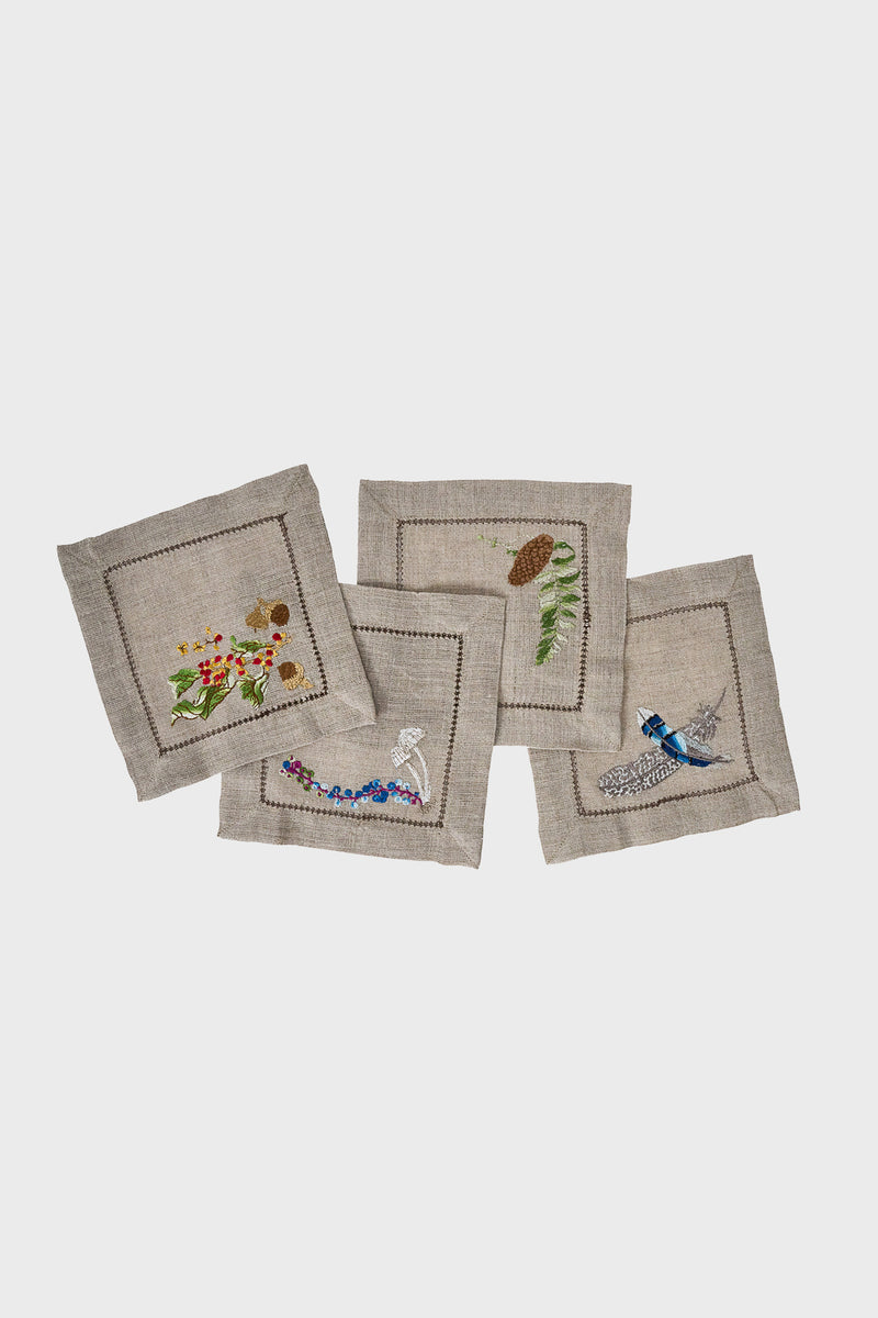 Set of four Juliska hand-embroidered linen cocktail napkins featuring nature-inspired designs—a bird, flowers, pinecone, and leaves—with elegant style.