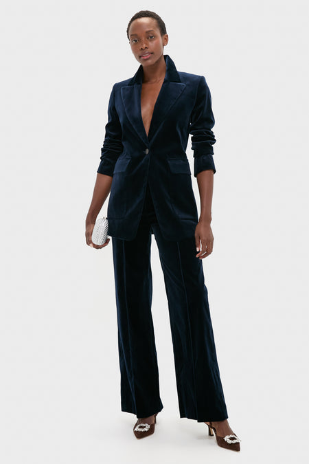 A person stands against a light gray background wearing Vanessa Bruno's cotton velvet tailored jacket, wide-leg pants, and single-button blazer, accessorized with a silver clutch and pointed heels.