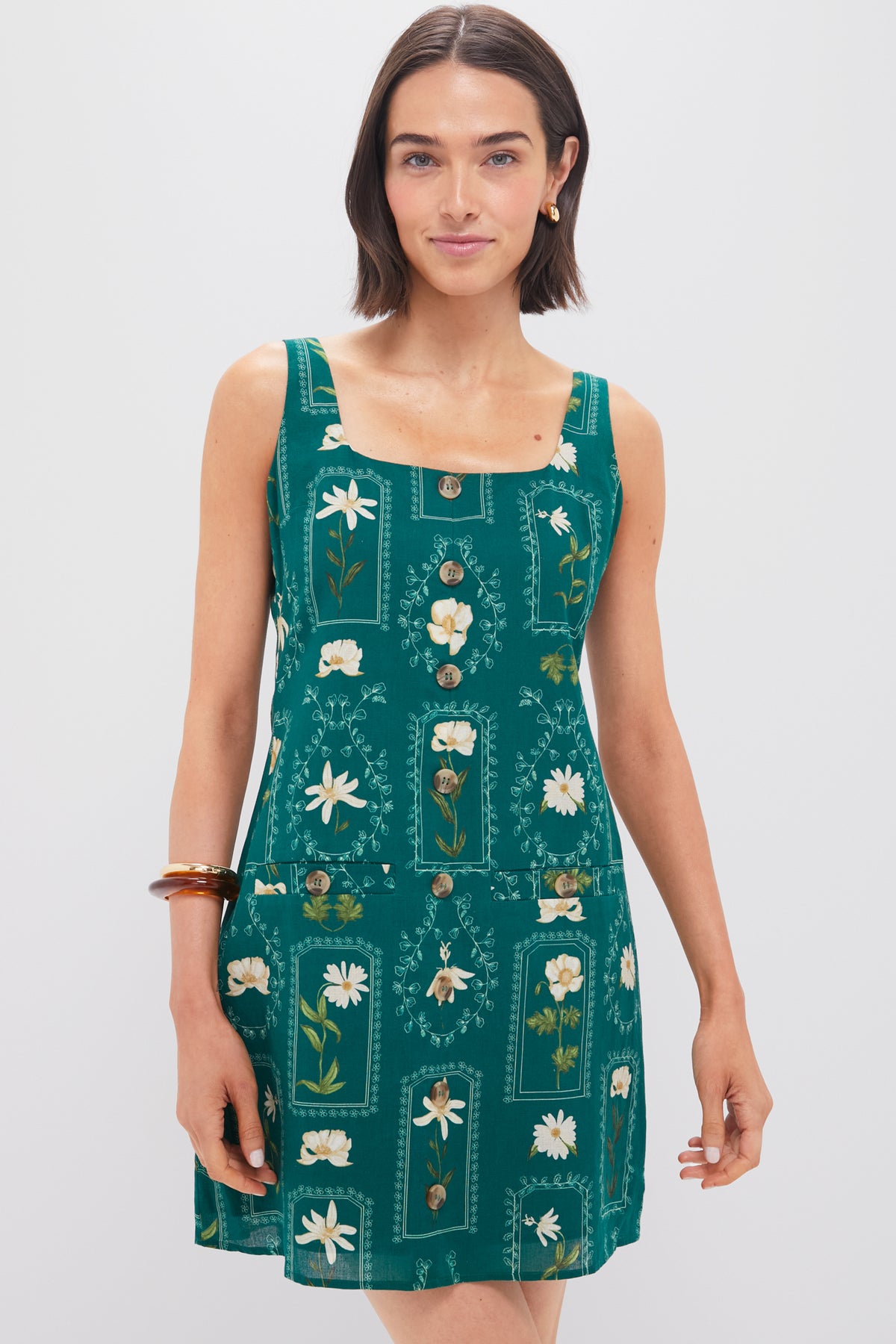 Woman wearing the Anna Cate Merrit Mini Dress, a sleeveless green dress with a square neckline, white floral print, and front buttons, posing tastefully.