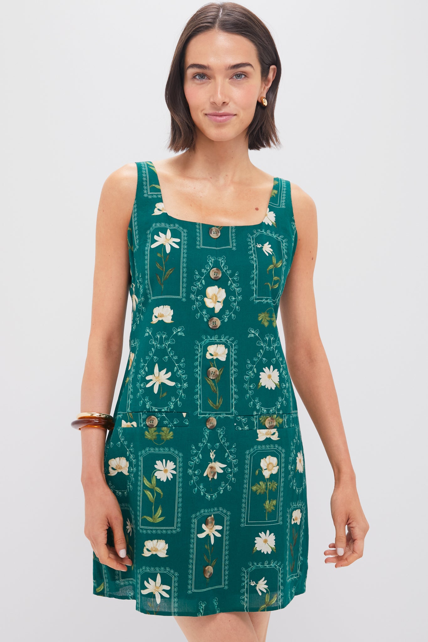 Woman wearing the Anna Cate Merrit Mini Dress, a sleeveless green dress with a square neckline, white floral print, and front buttons, posing tastefully.