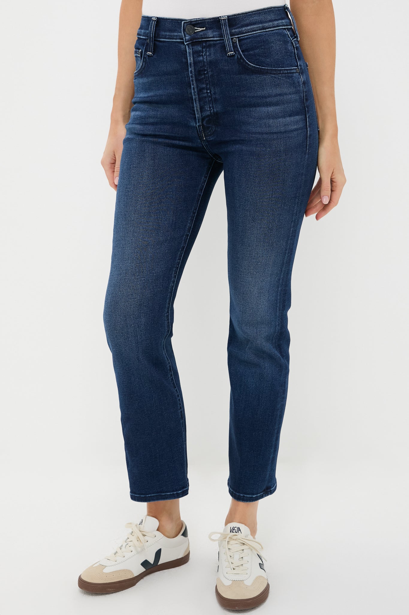Someone wears high-rise flared jeans in dark blue stretch denim by MOTHER, paired with white sneakers featuring beige and navy accents.
