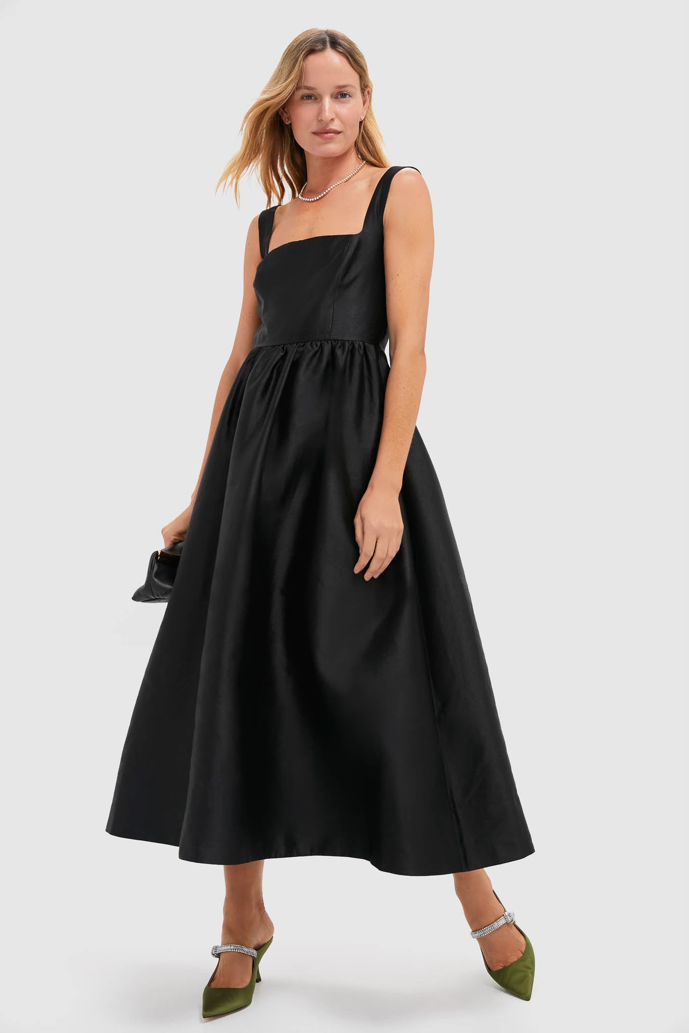 Woman in a classic black midi dress, showcasing Fern Satin Mae Heels—effortlessly chic olive-green heels with sparkly straps, embodying fun and high-end style.