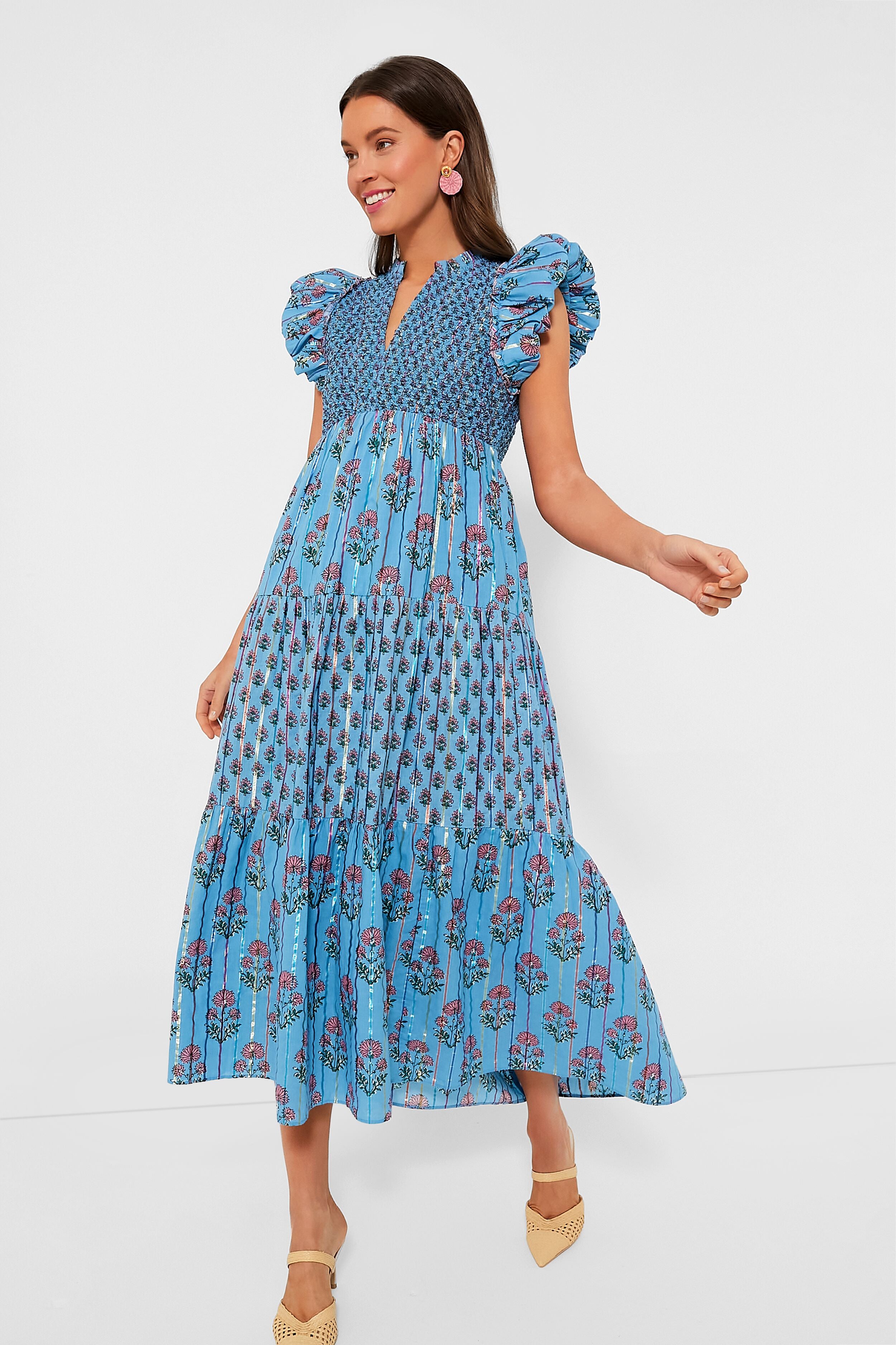 A woman wears a Saylor blue floral midi dress with puffed shoulders and a smocked bodice, paired with beige pointed-toe shoes and statement earrings.