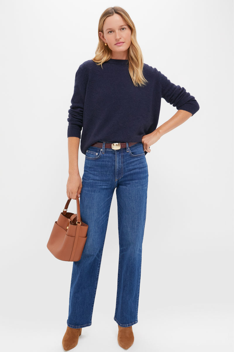 A woman stands against a white background wearing a Tuckernuck navy cashmere crewneck sweater, blue jeans, brown ankle boots, and holding a brown handbag.