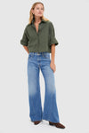Woman models the Duffle Bag Next Ex Shirt, a classic olive-green, boxy button-front with rolled cuffs, paired effortlessly with flared jeans and suede ankle boots.