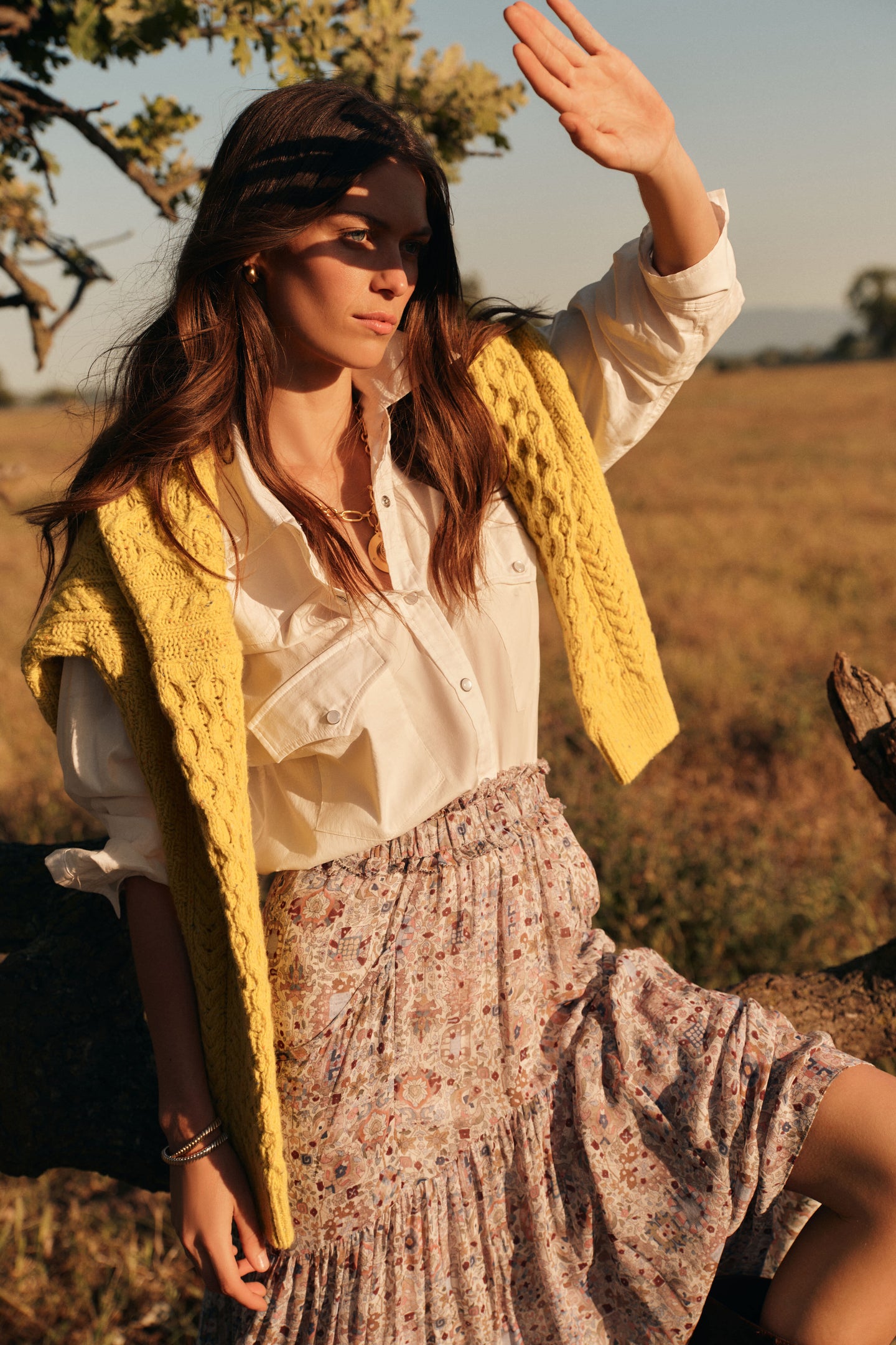 Woman standing outside in a long sleeved white button down shirt, a yellow cable knit sweater around her shoulders, a floral patterned maxi skirt, and chocolate brown boots. 
