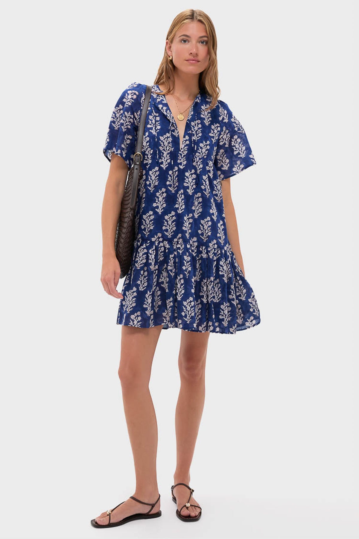 A woman stands against a plain background wearing a Love Tomas by Emerson Fry puff sleeve blue dress with a white leaf pattern, holding a black bag and wearing black sandals.