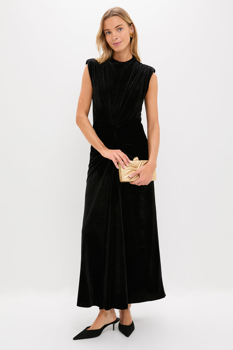 A woman in a Saylor sleeveless black floor-length dress, black pointed heels, and holding a gold clutch stands against a plain white background.