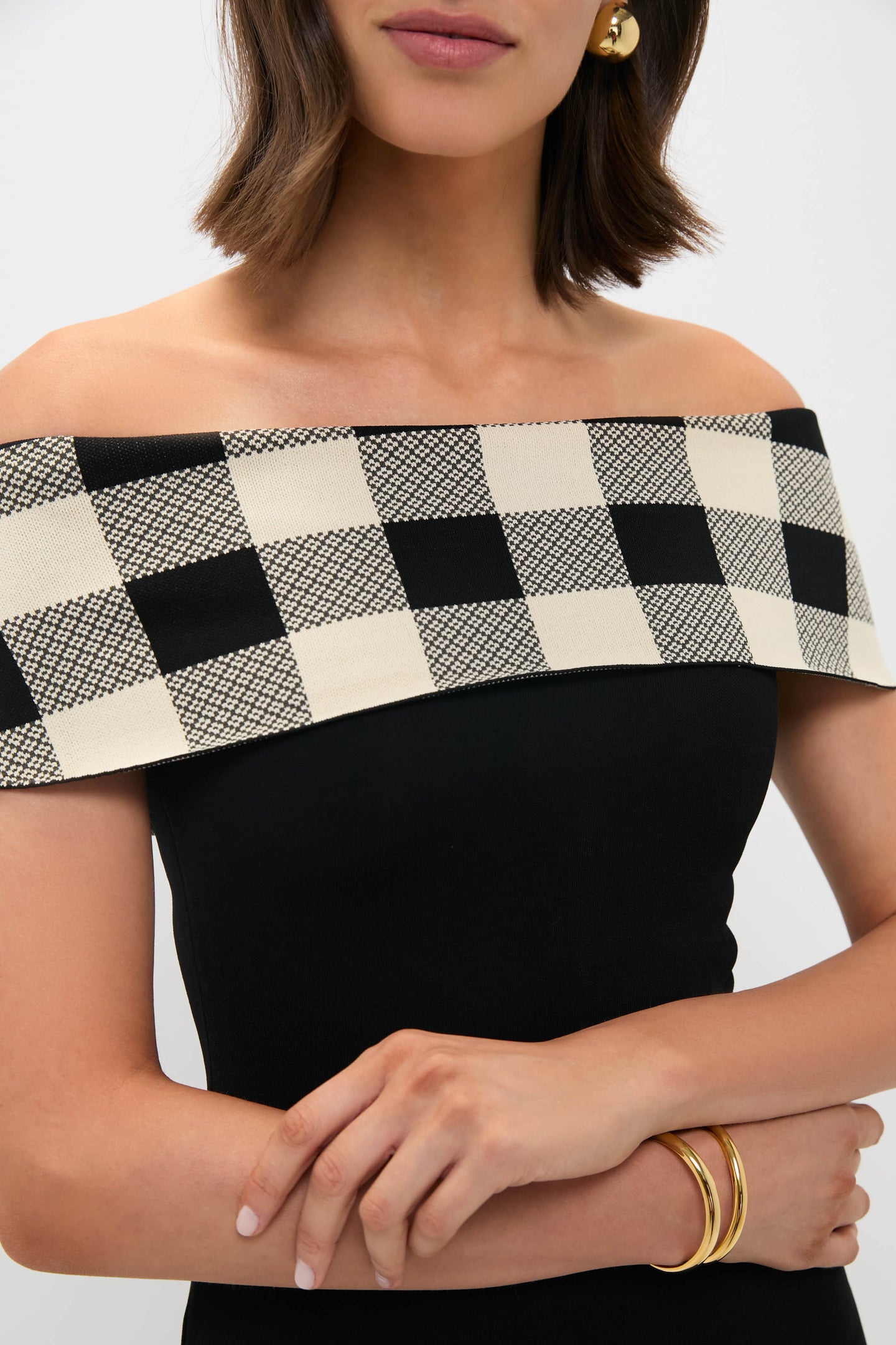 A woman in a POSSE black off-the-shoulder knit dress with a checkered beige and black neckline, gold hoop earrings, and two gold bangles stands with her arms crossed—an effortless nod to timeless women’s fashion.