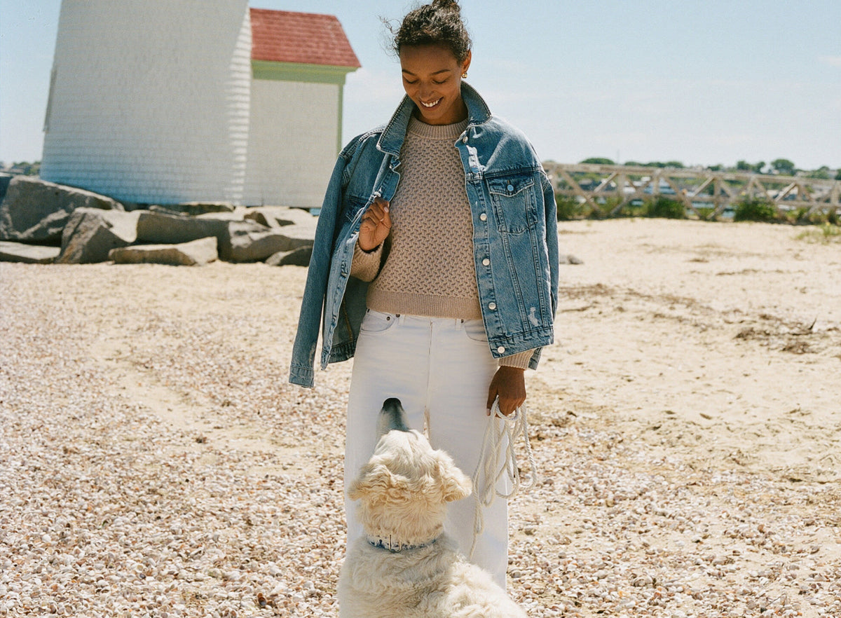 Woman in denim jacket with a tan sweater underneath wearing white jeans and holding a leash with a dog. 