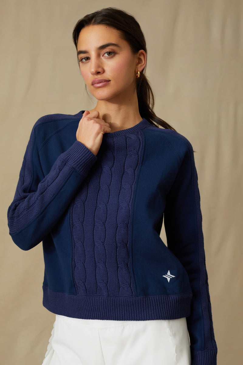 A woman poses confidently in a Tnuck Sport navy blue Cable Knit Sweatshirt with a small white logo on the hem and white pants.
