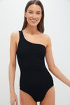 A woman smiles at the camera wearing a Hunza G black, high-cut crinkle swimsuit with a one-shoulder design.