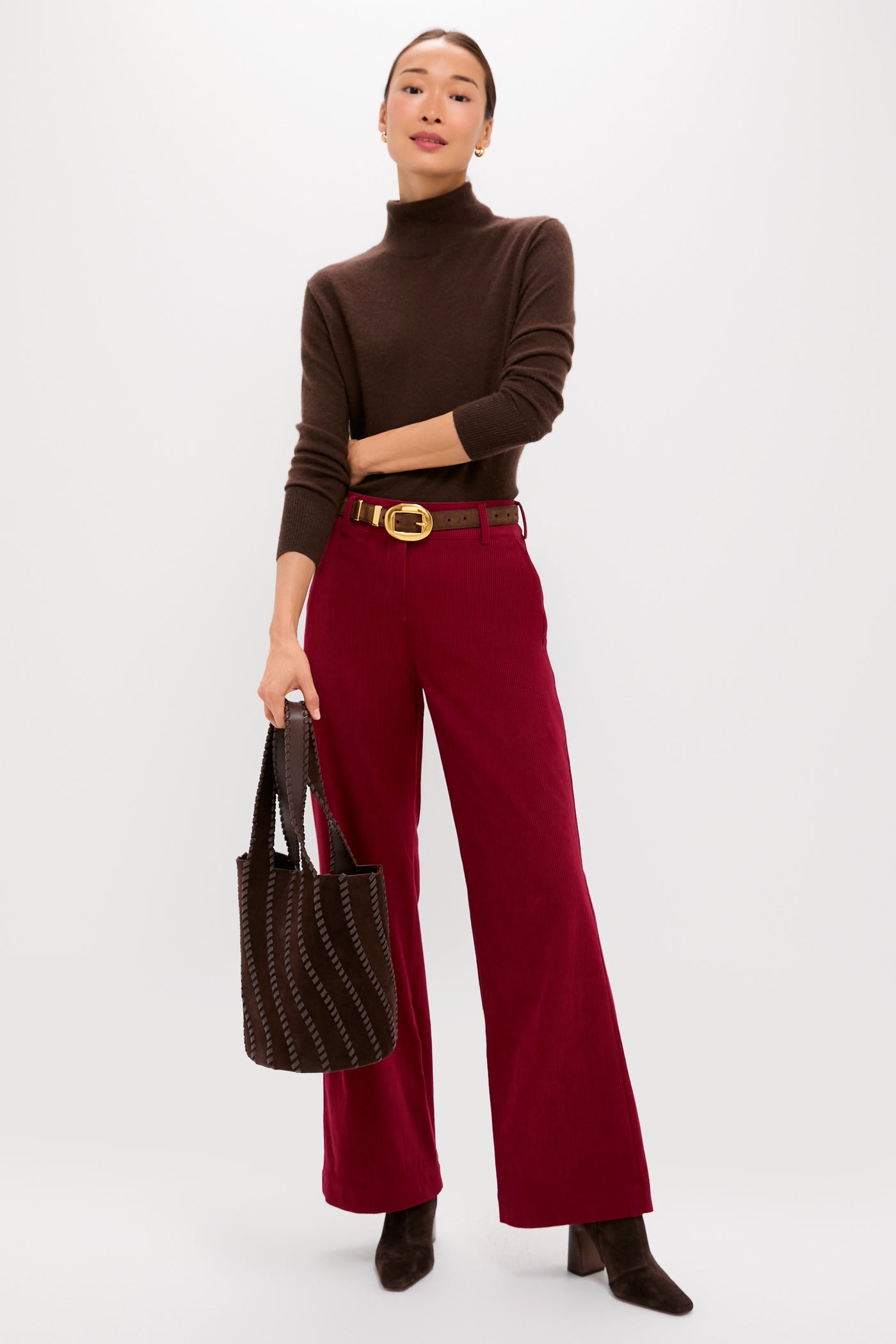 A woman wears a Tuckernuck brown turtleneck, wine wide-leg pants, brown boots, a gold-buckled belt, and holds a brown tote bag against a plain background.