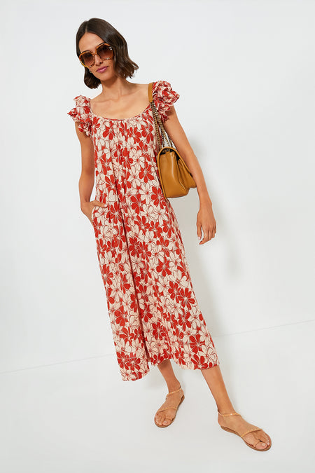 A woman wearing THE GREAT. red and white floral midi dress with ruffle sleeves, paired with tan sandals, a brown handbag, and sunglasses, stands against a plain white background.
